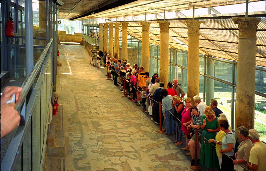 Il corridoio biabsidato nella struttura di Minissi, con i visitatori che camminano sopra i mosaici, protetti soltanto da un tappeto (foto di Jerzy Strzelecki - Opera propria, CC BY-SA 3.0, https://commons.wikimedia.org/w/index.php?curid=3208551)